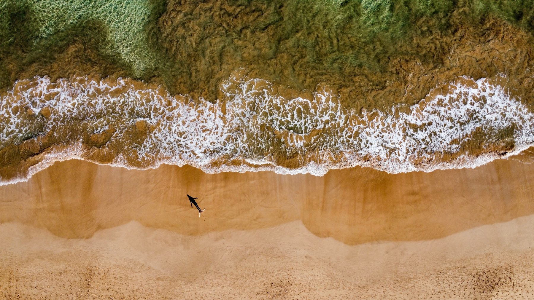 Aerial view of a person walking along a quiet sandy beach beside gentle waves